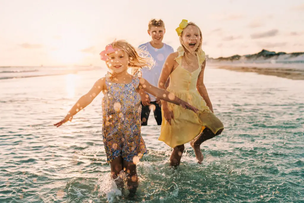 Three children running and splashing through shallow surf at sunset.
