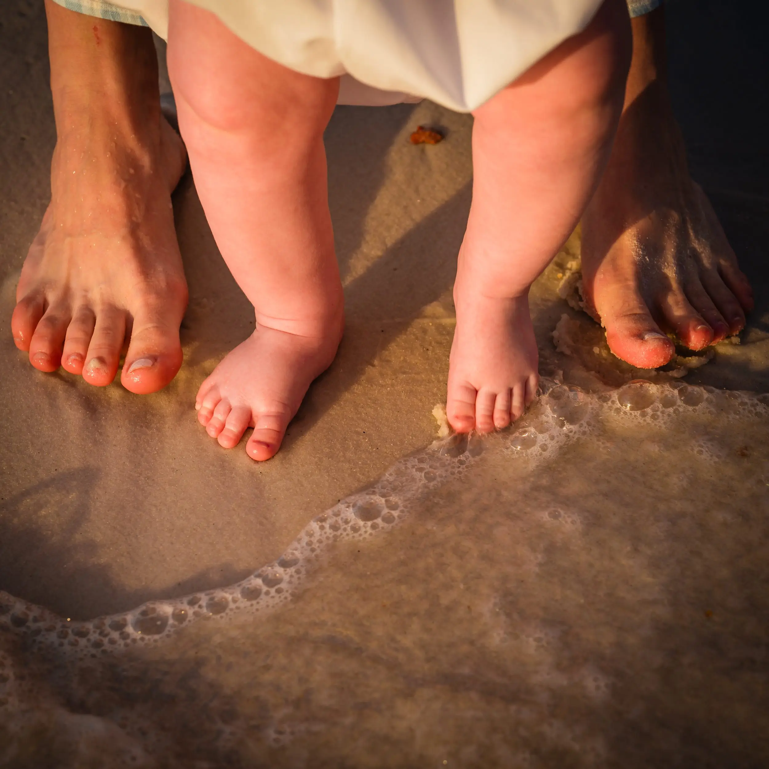 A baby's feet between an adult's feet at the water's edge.