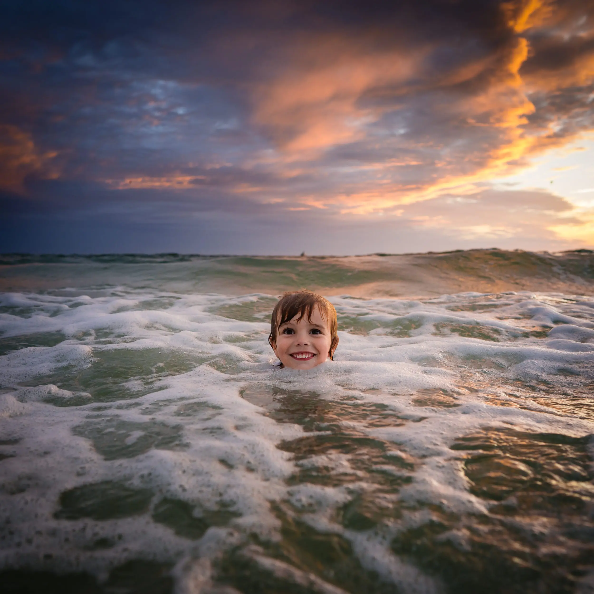 A smiling child in the foamy surf beneath a colorful sunset sky.