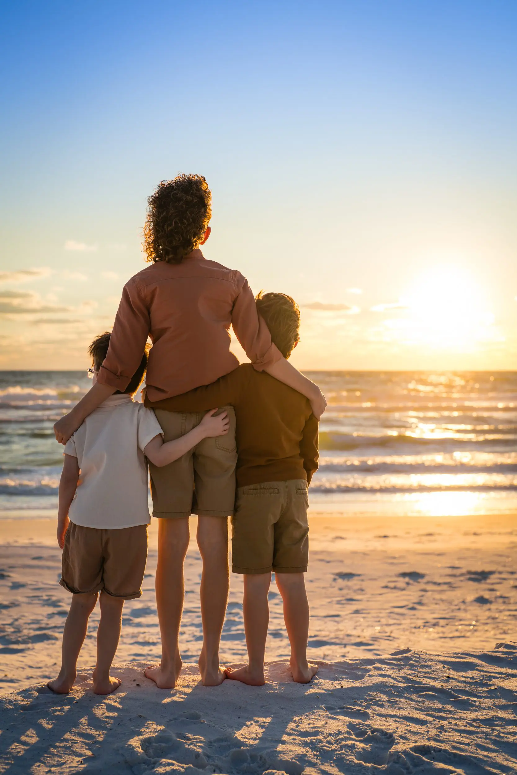 A parent and two children watching the sunset at the shoreline.
