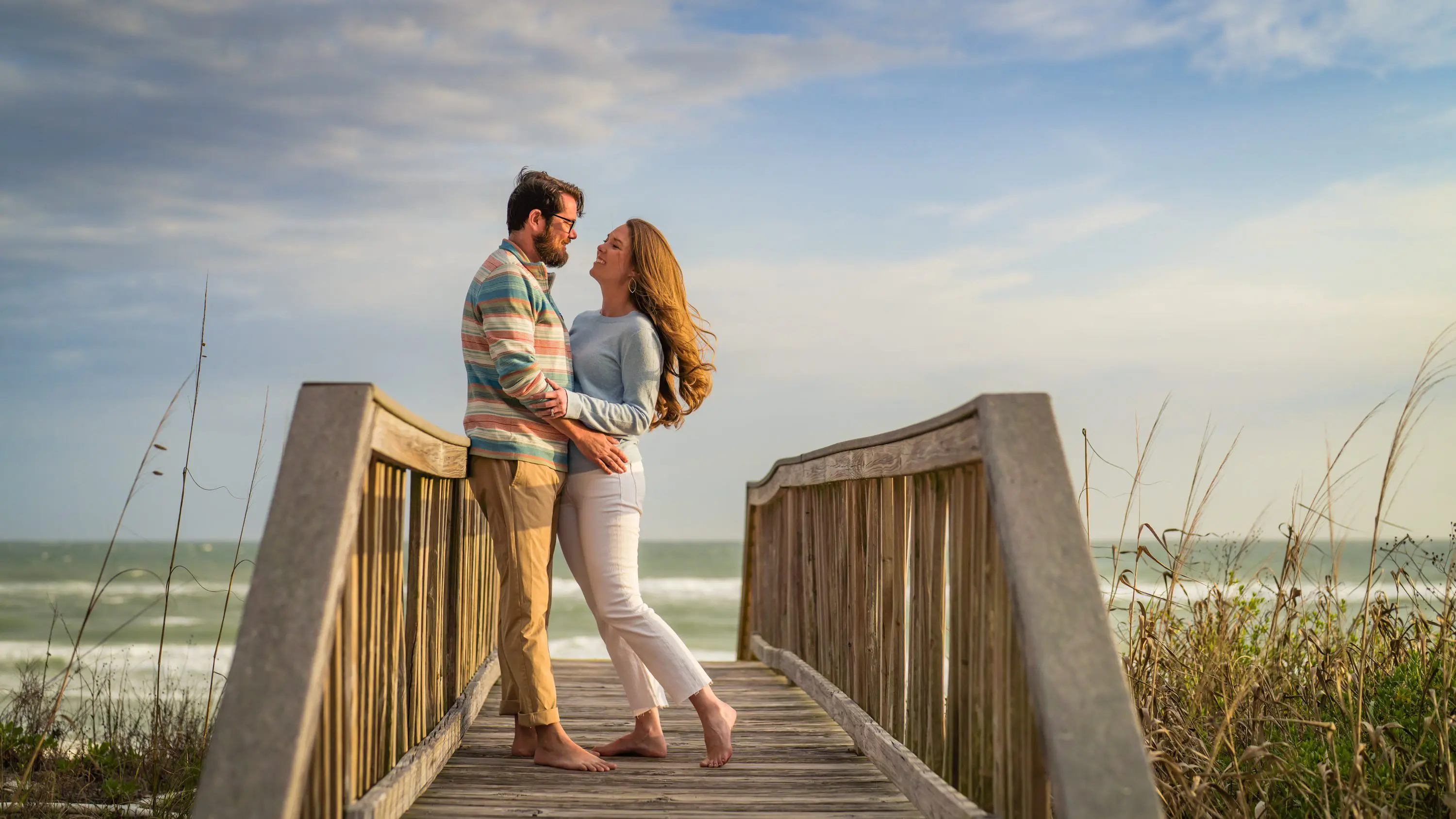 A couple embracing on a wooden beach boardwalk with the sea behind them.