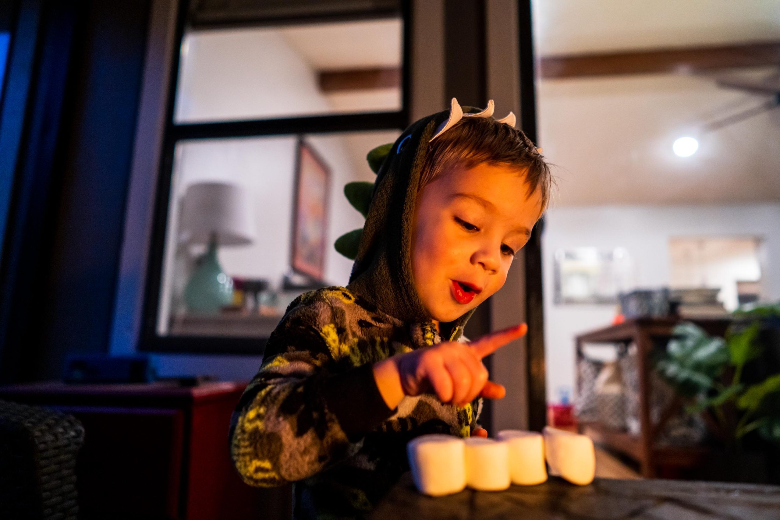 A child in dinosaur pajamas pointing toward marshmallows on a table.