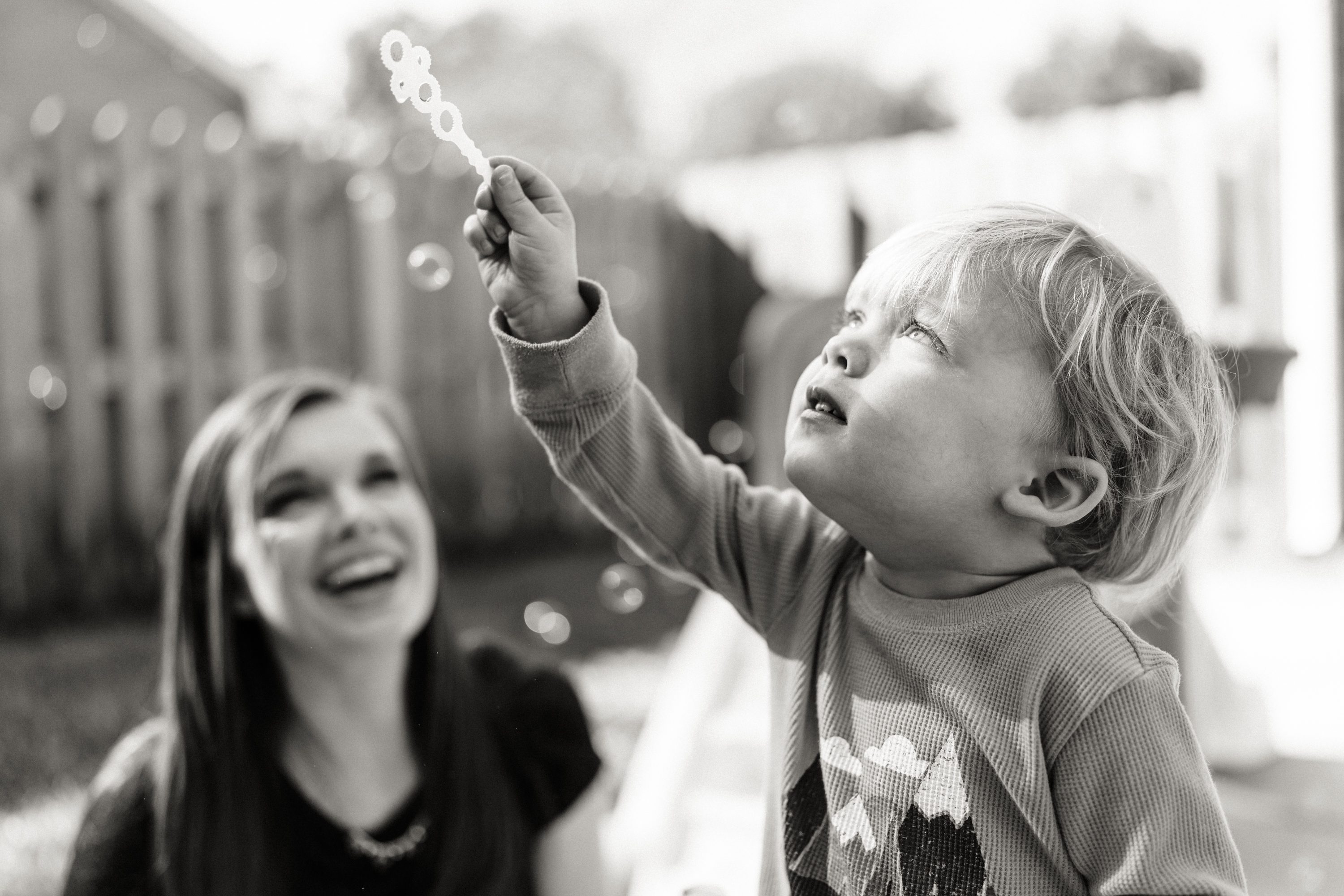 A young child blowing bubbles with an adult smiling in the background.