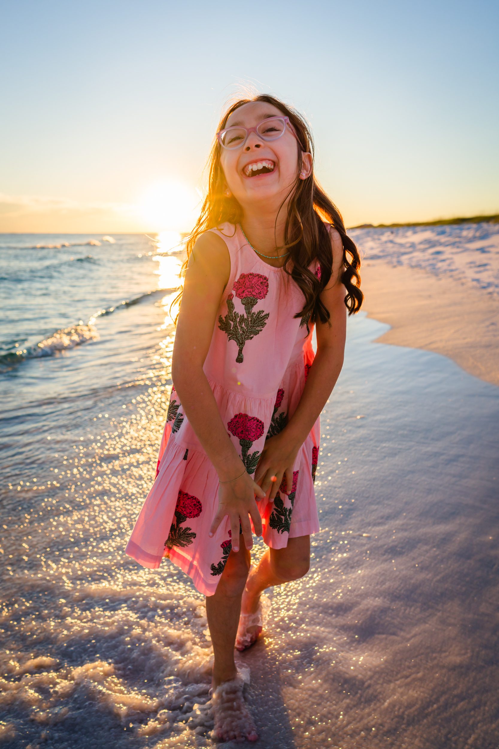 A girl laughing at the edge of the water in a pink dress.