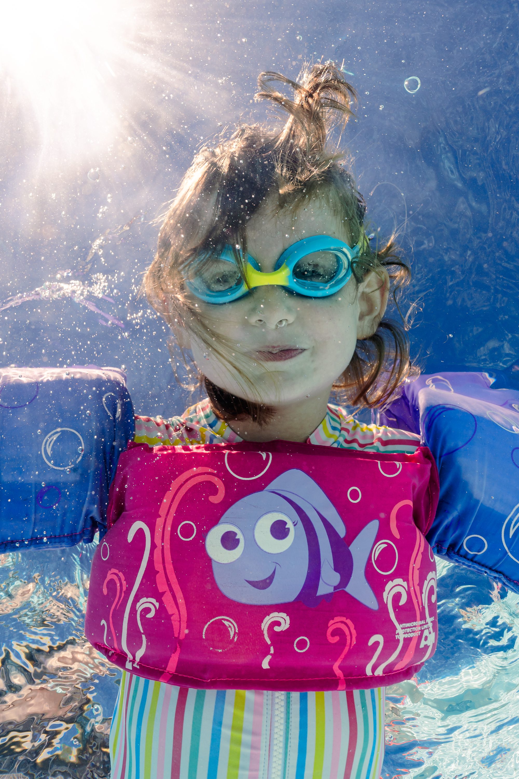 A child wearing swimming goggles and a pink flotation vest underwater.