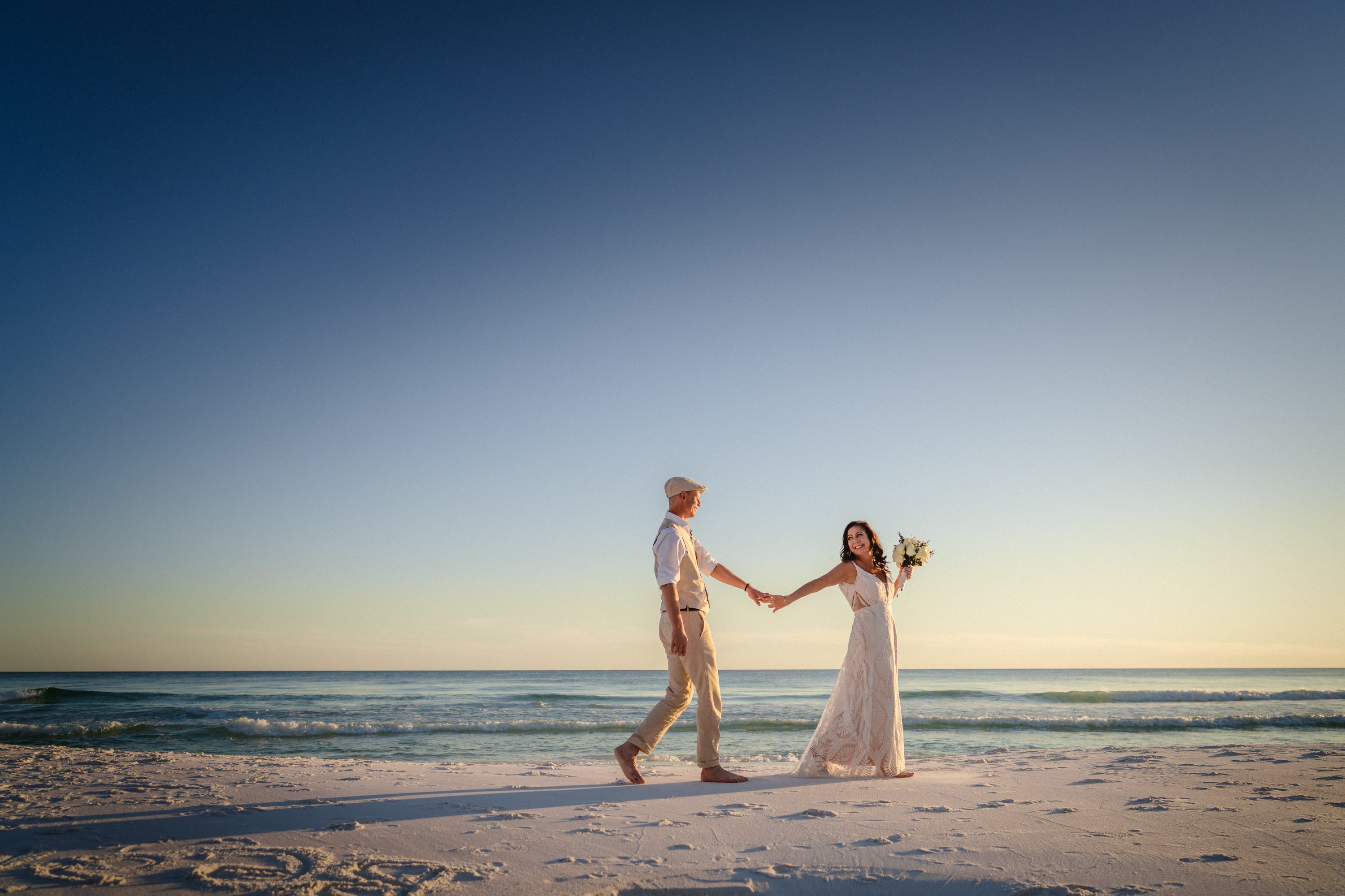 A bride holding a bouquet and her partner’s hand on the beach at sunset.