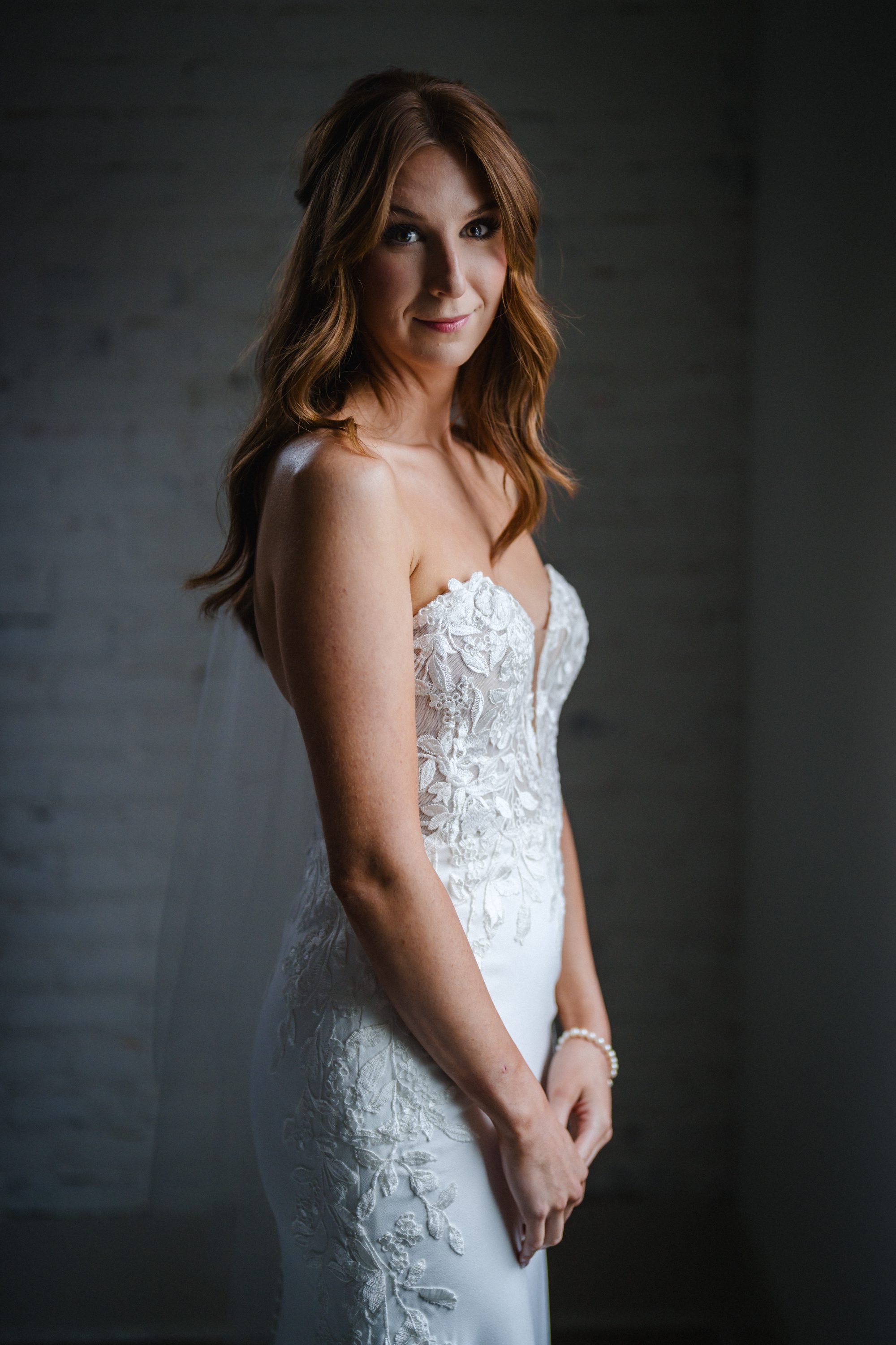 A bride in a lace wedding dress standing beside a dark wall.