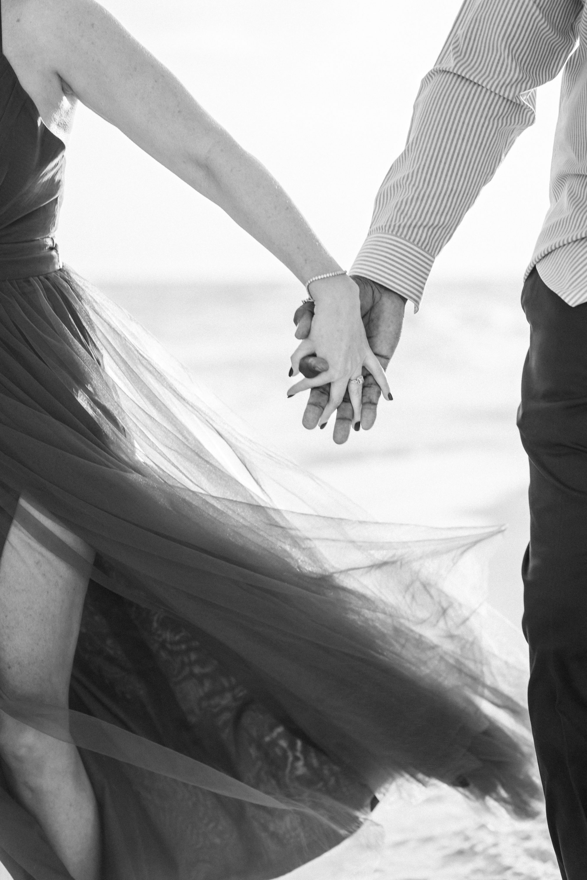 Black-and-white close-up of a couple holding hands on the beach, with a dress blowing in the breeze.