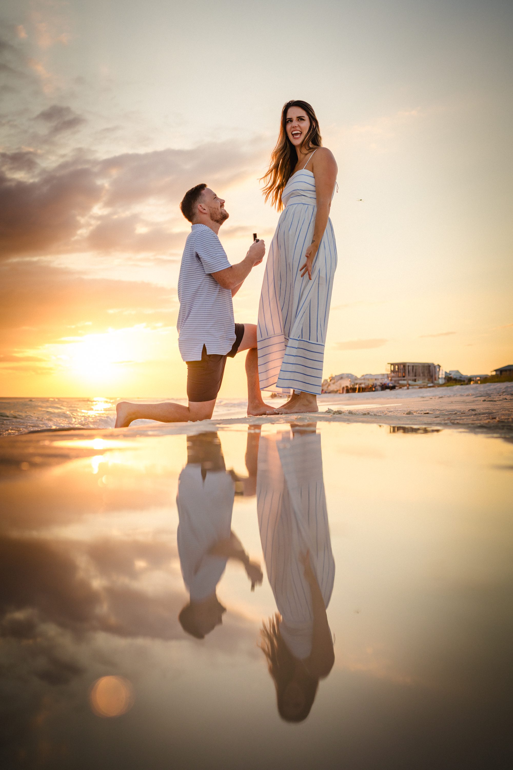 A man kneeling to propose on the beach at sunset, with the couple reflected in wet sand.