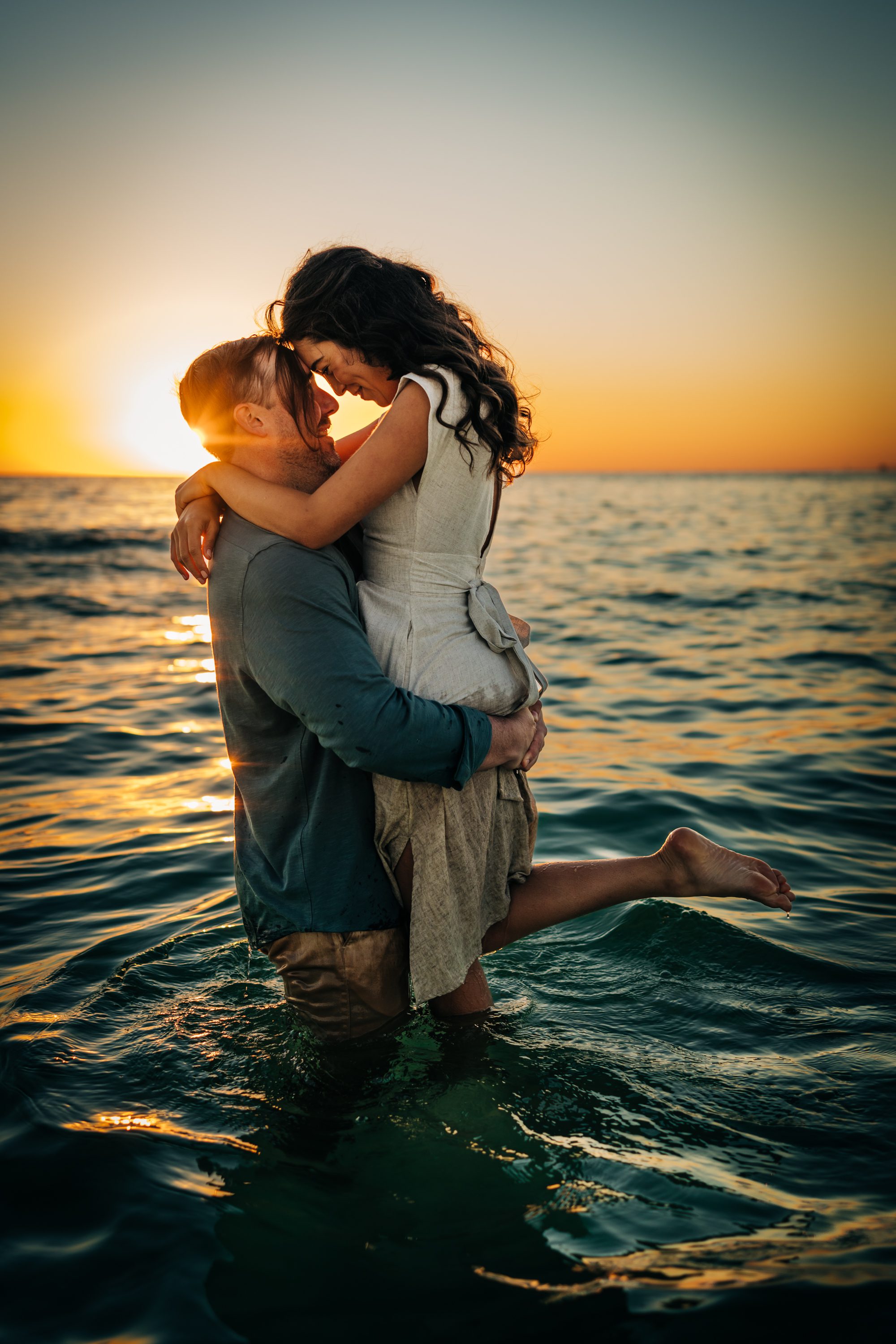 A couple embracing in the sea as one partner lifts the other at sunset.