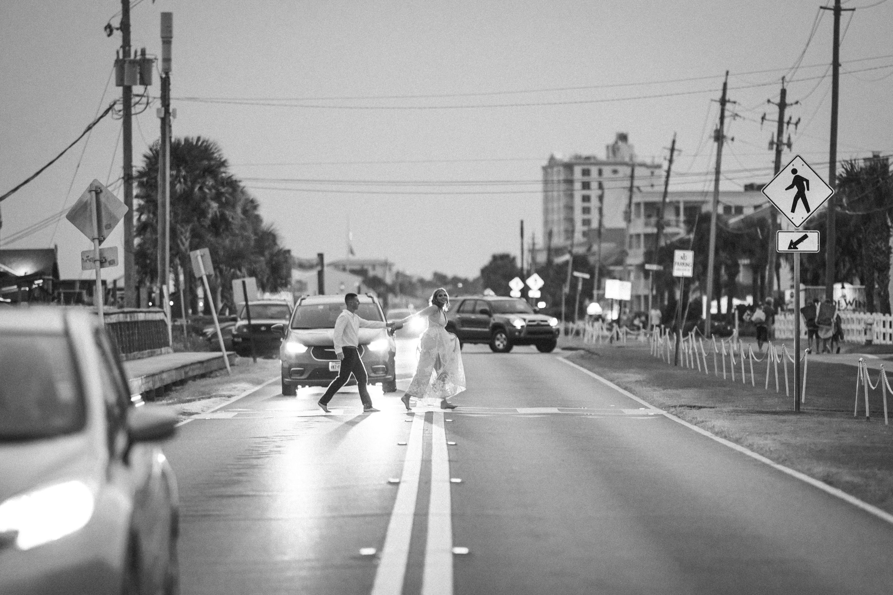 Black-and-white photograph of a couple holding hands while crossing a street at dusk.