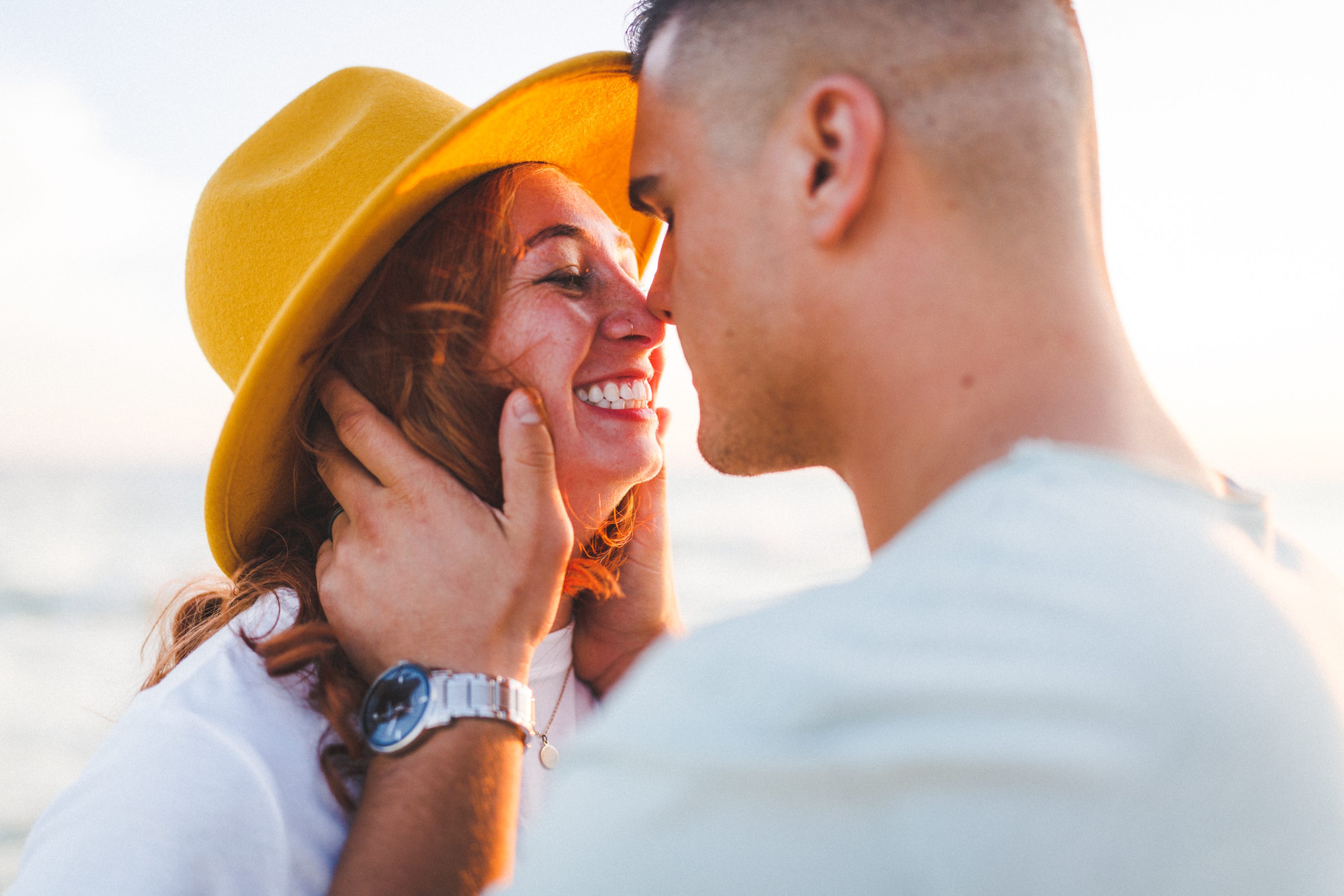 A smiling couple leaning close together, with one partner wearing a yellow hat.
