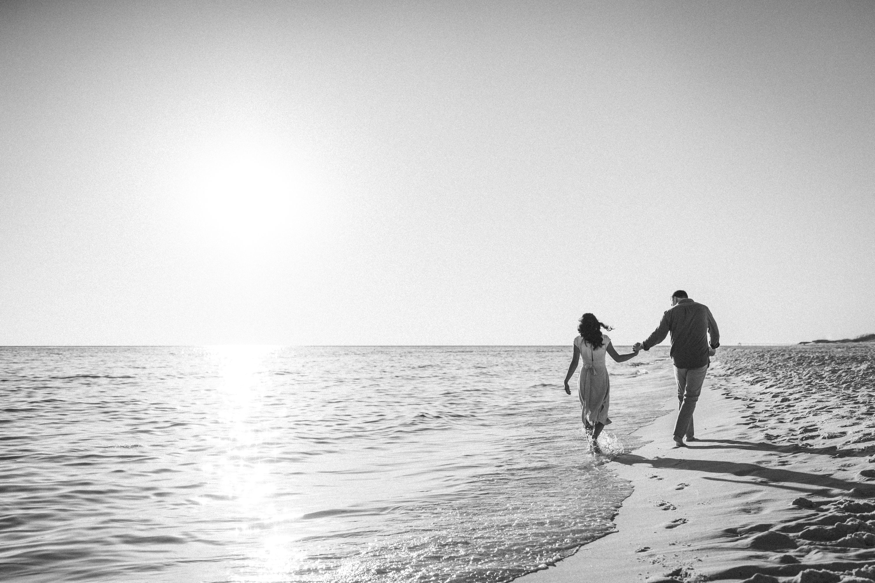 A couple holding hands as they walk along the shoreline in a black-and-white photograph.