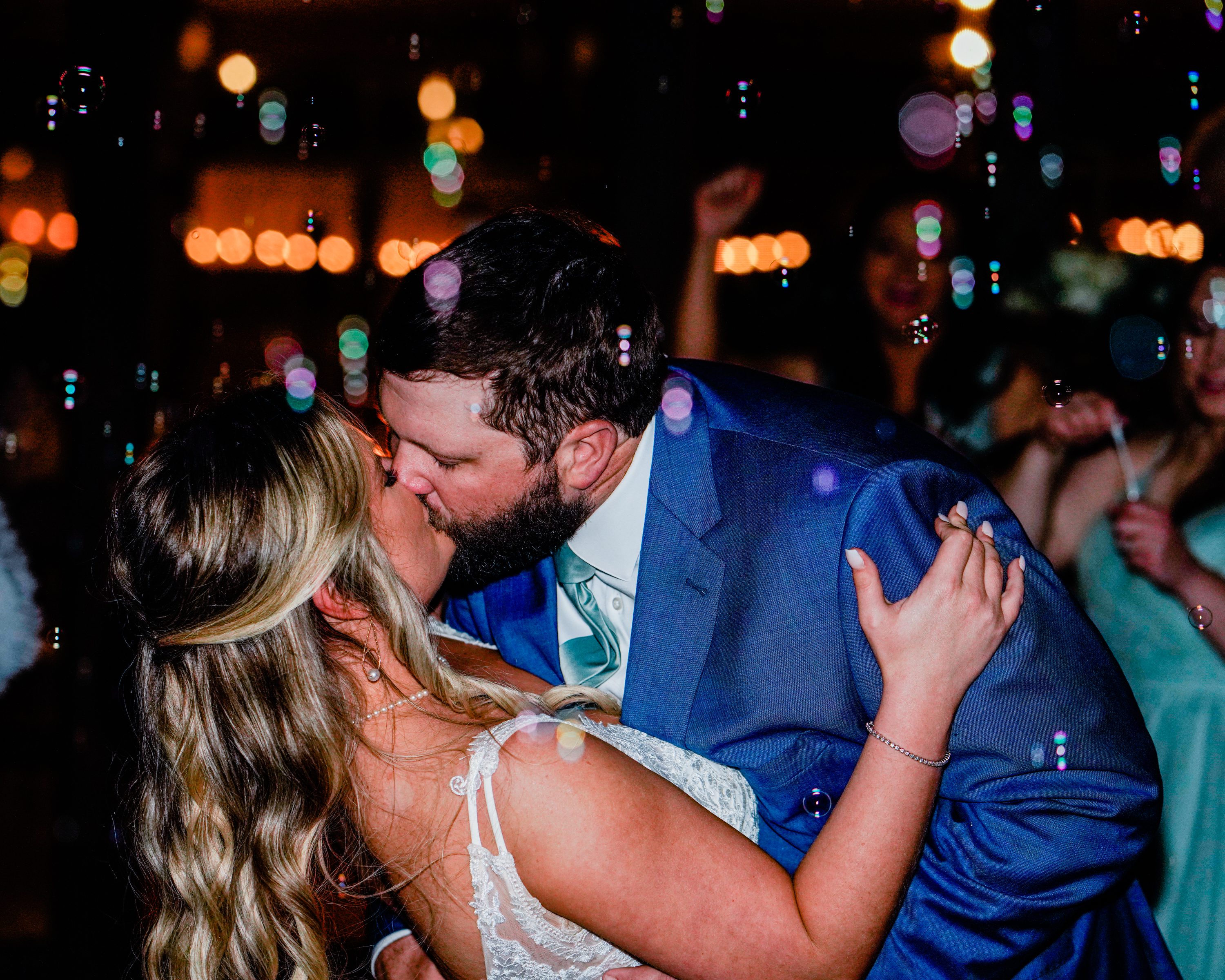 A bride and groom kissing on the dance floor beneath colorful reception lights.